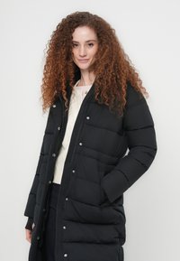 Μαύρο μπουφάν πάπλωμα με μακριά σιλουέτα, που διαθέτει φερμουάρ και κλείσιμο με κουμπιά, οριζόντιες ραφές και δύο πλαϊνές τσέπες.