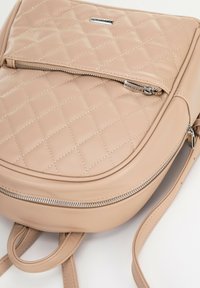 Sac à dos beige matelassé en cuir doux, avec une poche zippée à l'avant et des accents en matériel argenté, doté de bretelles plates pour un transport facile.