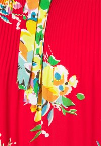 Röd tyg med ett blommönster i gult, grönt, blått och rosa. Har veck och en kontrasterande grön accent. Mjuk textur.