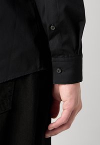 Main droite à côté d'un poignet de chemise noire à manches longues avec deux boutons, associée à un pantalon noir texturé sur un fond clair uni.