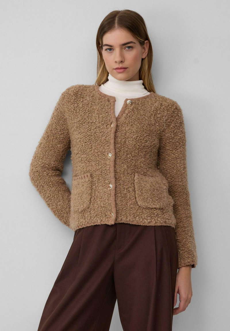 Vrouw in een beige gebreide knoop-om cardigan over een witte coltrui, gecombineerd met hoge donkere bruine wijde broek.