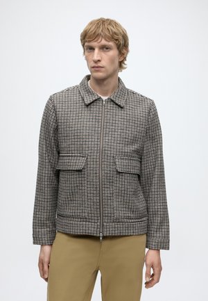 Blonde man wearing beige pants and a zippered gray and brown checkered jacket with two front flap pockets, standing against a plain white background.