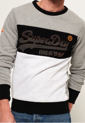 Mężczyzna ubrany w szarą, czarną i białą bluzę SuperDry z długimi rękawami i niebieskie dżinsy, stojący z jedną ręką w kieszeni.