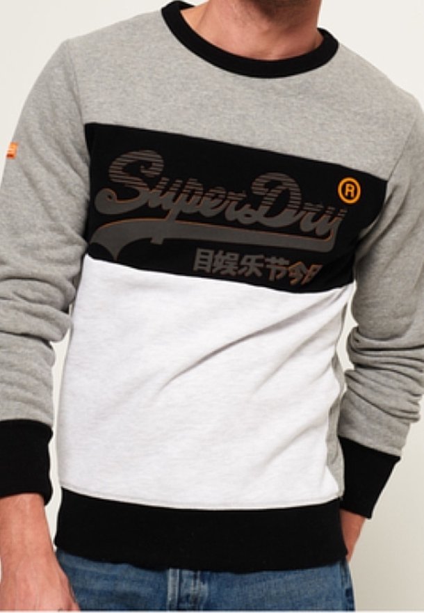Homme portant un sweat-shirt SuperDry gris, noir et blanc à manches longues et un jean bleu, debout avec une main dans la poche.