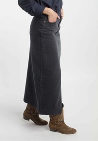 Jupe en denim noir longue avec poches, silouhette droite, tissu texturé et fentes latérales, assortie à des bottes de cow-boy brunes.