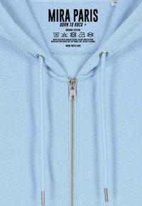 Hoodie zippé bleu clair avec fermeture éclair en métal et cordons, présentant un texte d'étiquette intérieure "MIRA PARIS BORN TO ROCK" et des instructions d'entretien.