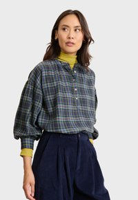 Camicia a quadri botton-up di colore blu scuro con accenti verdi e gialli, caratterizzata da maniche a sbuffo e colletto. Indossata sopra un dolcevita giallo.