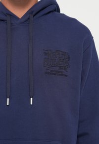 Navy blue hoodie with drawstrings and a black embroidered logo on the chest reading "The Replay Blue Jeans Authentic Premium Clothing."