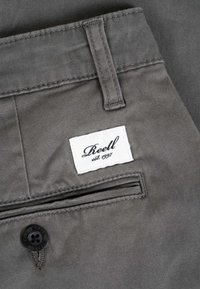 Poche arrière d'un pantalon gris avec fermeture boutonnée et étiquette blanche cousue au-dessus de la poche portant l'inscription « Reell est. 1997. »