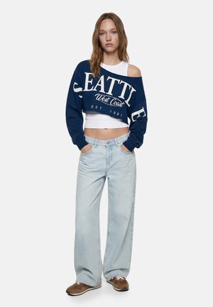 Jonge vrouw draagt een cropped marineblauwe trui met de opdruk "Seattle West Coast", een wit tanktopje, lichte wijdvallende spijkerbroek en bruine sneakers, handen in de zakken.