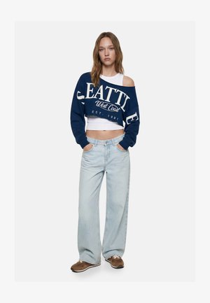 Jonge vrouw draagt een cropped marineblauwe trui met de opdruk "Seattle West Coast", een wit tanktopje, lichte wijdvallende spijkerbroek en bruine sneakers, handen in de zakken.