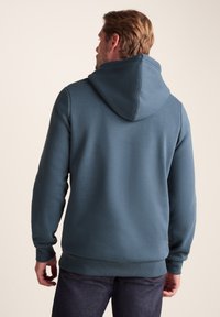 Hooded sweatshirt in a muted blue, featuring a textured fabric, ribbed cuffs, and a hem. Back view shows a relaxed fit and adjustable hood.