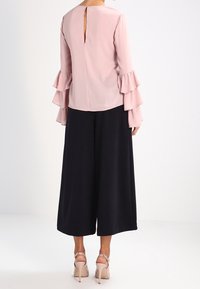 Blusa rosa con maniche a volant, dettaglio a chiave sulla schiena, abbinata a pantaloni neri a gamba larga. Le scarpe sono di un rosa chiaro con cinturini alla caviglia.