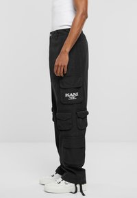 Svarte cargo-bukser med flere cargo-lommer, løs passform og KANI-logo detalj. Stoffet er mykt med synlig søm. Hvite joggesko er brukt.