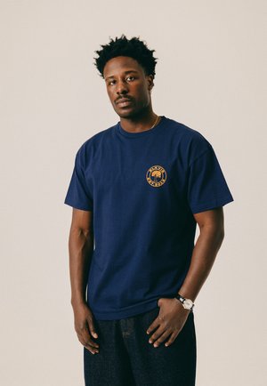 Mand med kort krøllet hår iført en marineblå T-shirt med et gult rundt trælogo og mørke jeans, stående mod en beige baggrund.