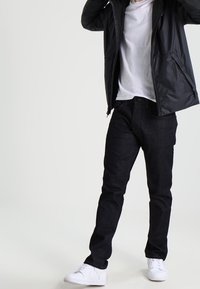 Svart lättviktjacka, vit crew neck t-shirt, slim-fit mörka denimjeans och vita sneakers. Avslappnat outfit med rena linjer och neutrala färger.