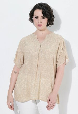 Femme aux cheveux courts et foncés portant un chemisier beige à fleurs et un pantalon blanc, debout devant un fond blanc avec un petit tatouage de cœur sur le bras.
