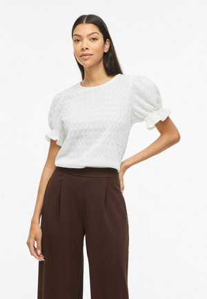 Kobieta stoi z ręką na biodrze, ubrana w białą teksturowaną bluzkę z bufkami oraz wysokie brązowe spodnie o szerokich nogawkach.