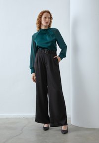 Blouse en satin bleu canard avec un col haut et des détails plissés, associée à un pantalon large taille haute noir et des talons noirs.