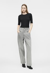 Femme aux cheveux longs portant un haut noir ajusté, un jean gris ample avec les mains dans les poches, et des bottines noires, se tenant devant un fond blanc.