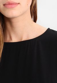 Blusa nera con scollatura ampia e arrotondata, tessuto morbido con finitura opaca e delicati dettagli arricciati nella scollatura.
