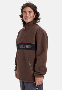 Sudadera de forro polar marrón con cuello alto, cierre de media cremallera en la parte delantera, y una banda horizontal negra con logo blanco. Incluye bolsillos con cremallera y detalles en rojo.