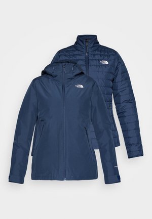 Temno modra zimska jakna s kapuco in zadrgo. Ima prešito teksturo, stranske žepke in logo na prsih. Obrnljiva zasnova.