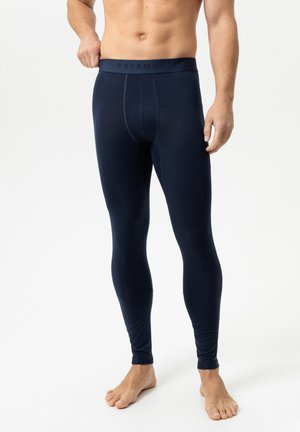 Personne torse nu portant des leggings thermiques bleu foncé ajustés, debout pieds nus sur un fond blanc.