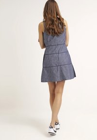 Vestido de mezclilla con un torso ajustado y falda acampanada, en azul oscuro, con costuras decorativas. Combinado con zapatillas negras y blancas.