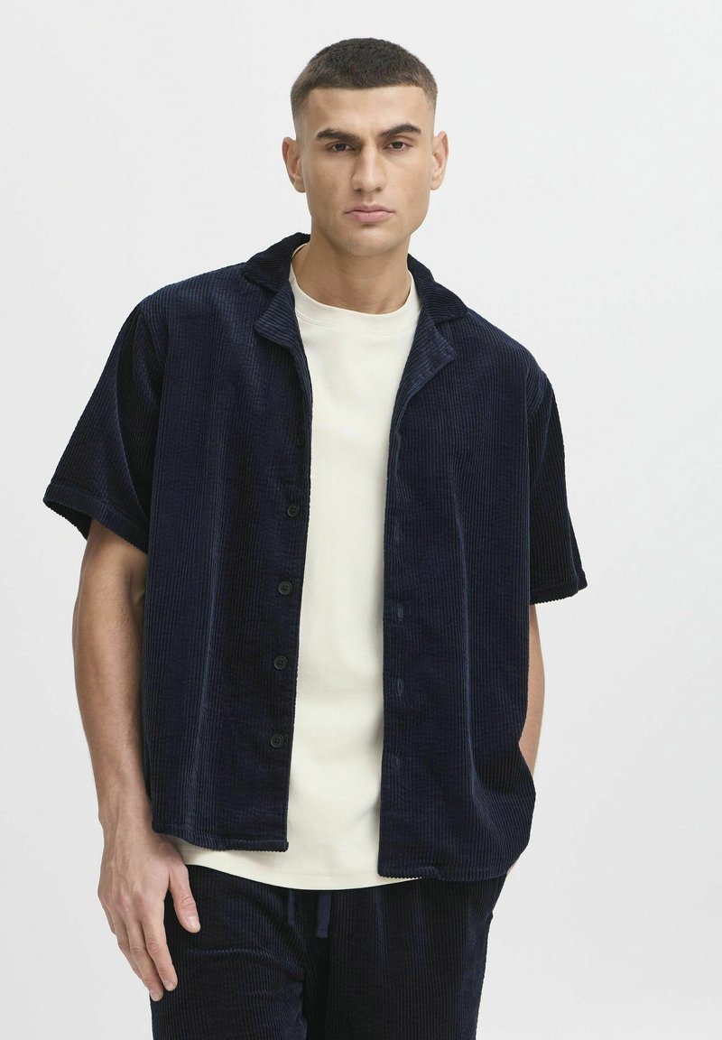 Chemise courte à manches en velours côtelé bleu marine, avec un col en V et des boutons sur le devant, portée par-dessus un t-shirt crème clair. Design simple et structuré.