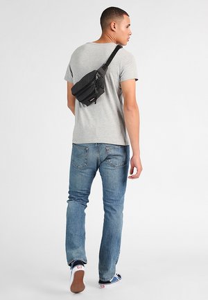 Black fanny pack with two zippered compartments, adjustable strap, and textured surface worn over a gray t-shirt, paired with blue jeans and sneakers.