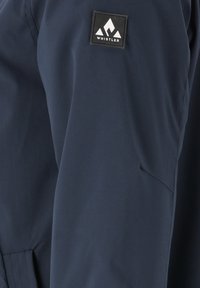 Chaqueta azul marino con tejido liso y resistente al agua. Incluye un parche cuadrado negro con un diseño de montaña blanca en la parte superior del brazo.
