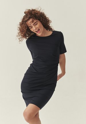 Młoda kobieta o kręconych włosach, ubrana w dopasowaną czarną sukienkę, lekko się pochylająca, z zamkniętymi oczami i uśmiechem z otwartymi ustami na jednolitym tle.