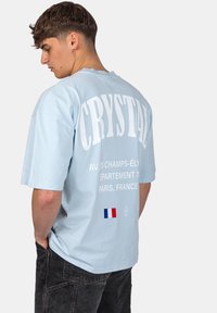 Tyrkysové tričko s krátkým rukávem, na zádech s výrazným bílým textem, obsahující detail francouzské vlajky a diamantové logo. Měkký materiál s volným střihem.