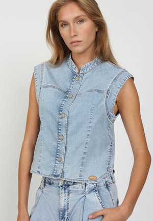 Junge Frau trägt ärmellose hellblaue Jeansweste mit Knöpfen und passende hoch taillierte Jeans, neutraler Gesichtsausdruck, einfarbiger Hintergrund.