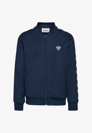 Kind steht und trägt einen marineblauen Trainingsanzug mit weißen Chevron-Streifen an Ärmeln und Beinen, weiße Sneaker mit grünen Akzenten.