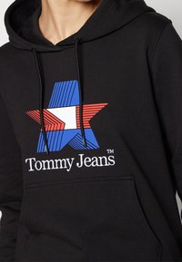 Černá mikina s kapucí, která obsahuje velký vícolorový grafiku hvězdy v modré, červené a bílé barvě, s logem "Tommy Jeans" vyšitým bílou nití.