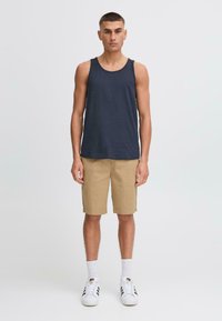 Marineblå tanktop, beige shorts, hvide sokker og sorte sneakers med accenter. Modellen står oprejst med et neutralt udtryk foran en lys baggrund.