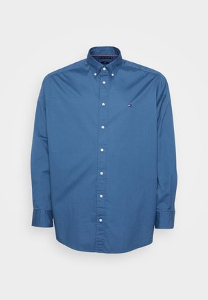 Sininen nappikauluspaita puuvillasta, jossa on terävä kaulus, pitkät hihat ja nappi-olkain. Vasemmassa rinnassa pieni logo.