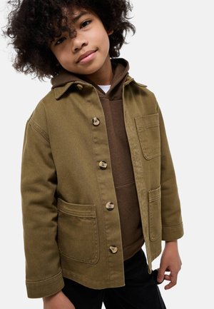 Garçon aux cheveux bouclés portant une veste marron à boutons par-dessus un sweat à capuche marron, debout avec un léger sourire sur un fond blanc uni.