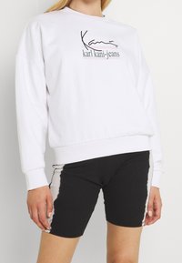 Sudadera blanca con el logo "karl kani-jeans" en negro, con un corte relajado y puños de canalé, combinada con pantalones cortos de ciclista negros con un detalle degradado.