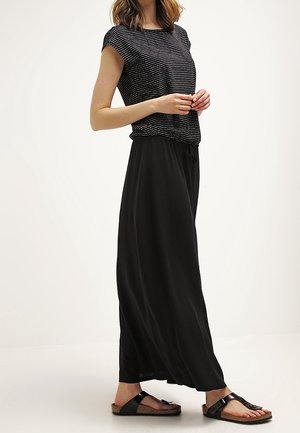 Femme portant un haut à manches courtes rayé noir et blanc, un pantalon large noir ample et des sandales noires, debout devant un fond blanc.