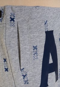 Pantalones de chándal grises con una gran "A" azul oscura, costuras abstractas en azul y bolsillos laterales con una textura suave.
