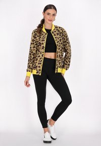 Gul och svart leopardmönstrad bomberjacka med ribbade ärmslut, bärs över en svart crop top och svarta leggings, i kombination med vita sneakers.