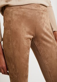 Los pantalones de ante marrón presentan una textura suave, un corte ajustado y detalles cosidos a lo largo de los lados. La tela parece suave y acolchada.