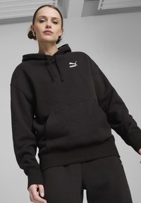 Černá mikina s kapucí, která má klokanní kapsu, žebrované manžety a lem. Na hrudi se nachází bílé logo Puma. Měkká, strukturovaná látka.