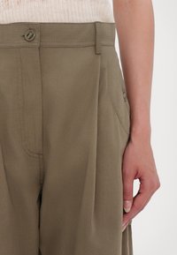 Gros plan d'une personne portant un pantalon kaki taille haute avec un bouton et une poche latérale, et un haut blanc côtelé.