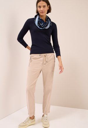 Beige Jogginghosen mit Kordelzug und seitlichen Reißverschlüssen, kombiniert mit einem taillierten navy-blauen Langarmshirt und einem gemusterten blauen Schal. Sneakers sind sichtbar.