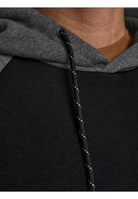 Inquadratura ravvicinata di una persona che indossa un maglione nero con cappuccio grigio e cordino nero con macchie bianche sopra una maglietta marrone.