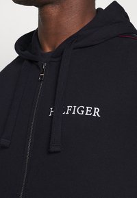 Sudadera con cremallera de marina, mezcla de algodón, que presenta el logo blanco "HILFIGER", capucha con cordón y tejido texturizado con un sutil acento rojo en el hombro.